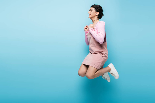 full length view of excited woman with clenched fists levitating on blue background - Photo, Image