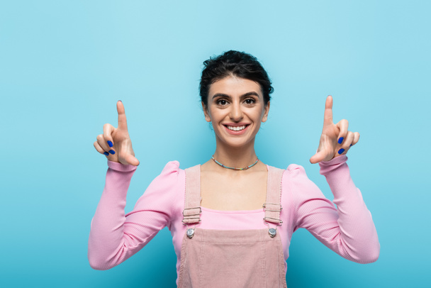 front view of cheerful and stylish woman pointing up with fingers isolated on blue - Foto, afbeelding