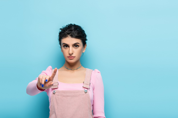 young brunette woman pointing with finger and looking at camera on blue background - Foto, afbeelding