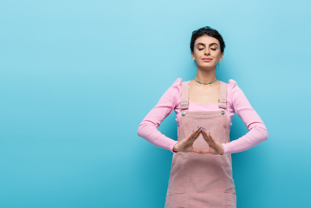 positive woman in pastel clothes relaxing in meditation pose isolated on blue - Zdjęcie, obraz