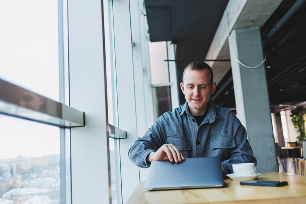 Ein junger erfolgreicher Geschäftsmann im Anzug trinkt Kaffee und arbeitet in einem Café mit drahtlosem, kostenlosem Internet auf einem Laptop und sitzt allein an einem Tisch in einem Café. - Foto, Bild
