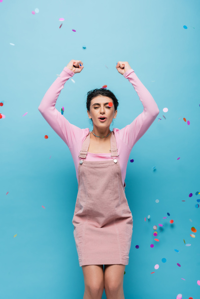 excited woman with closed eyes showing triumph gesture under falling confetti on blue background - Φωτογραφία, εικόνα