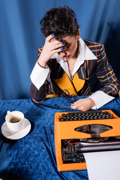 exhausted journalist holding eyeglasses near vintage typewriter and coffee cup on blue background - Photo, Image