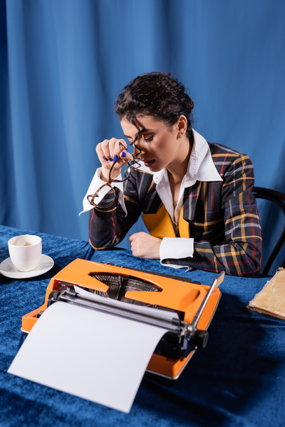 periodista cansado sentado con los ojos cerrados cerca de taza de café y máquina de escribir vintage sobre fondo azul - Foto, Imagen