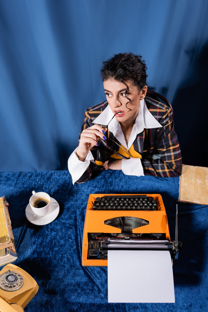 vista de alto ângulo de estilo vintage newswoman perto de máquina de escrever com papel em branco na toalha de mesa de veludo azul - Foto, Imagem