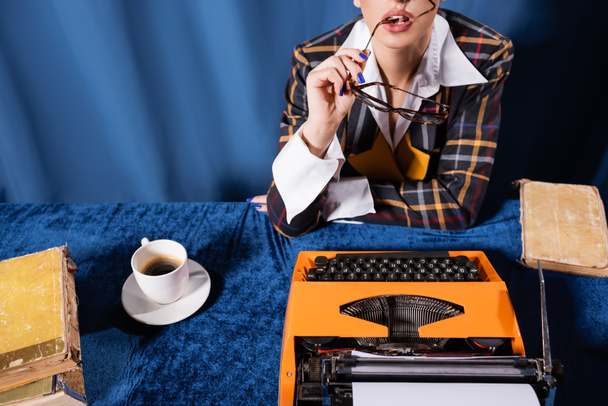 partial view of vintage style woman holding eyeglasses near typewriter and coffee cup on blue background - Foto, afbeelding