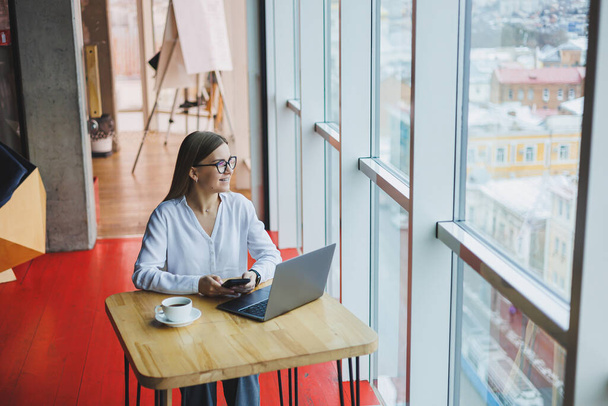 Egy okos nő menedzser egy csésze kávéval üzleti beszélgetést folytat egy okostelefonon keresztül, hogy megvitassák a szállítási megbízást, egy fiatal nő kis tulajdonosa ül egy asztalnál egy kávézóban egy laptoppal és felhívja - Fotó, kép