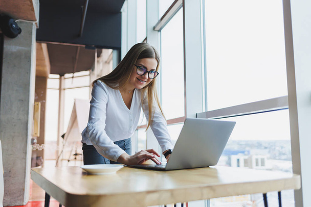 Aantrekkelijke blonde vrouw in casual kleding en bril concentreert en typt op een laptop terwijl ze aan een tafel zit in een café met koffie, een studentmeisje in een wit shirt en een bril - Foto, afbeelding