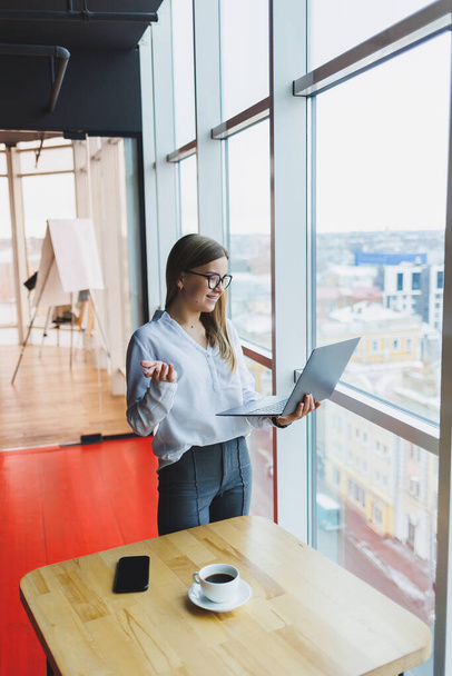 Iloinen nuori vaalea liiketoiminnan nainen Euroopan ulkonäkö valkoinen paita ja lasit pitää kannettavan kannettavan tietokoneen käsissään, seisoo lähellä suuri ikkuna toimistossa ylimmässä kerroksessa - Valokuva, kuva