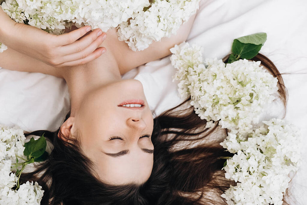 Charming young brunette girl 20s old years with perfect makeup. Close up portrait of pretty cute caucasian woman lying on comfy white bed with fresh spring beautiful flowers. Skincare concept - Фото, зображення