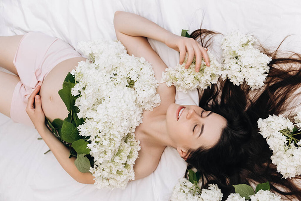 Portrait of beautiful cute smiling young pregnant woman with eyes closed lying on comfy bed with fresh white flower, enjoys wonderful morning and pregnancy. Childhood and future mom concept - Fotografie, Obrázek