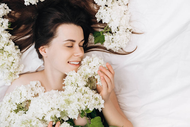Close up portrait of cute beautiful young female 20s with closed eyes and sincere smile lying in cozy white bed, spring flowers around head. Relaxed millennial lady with clear skin enjoys, feels joy - Fotografie, Obrázek