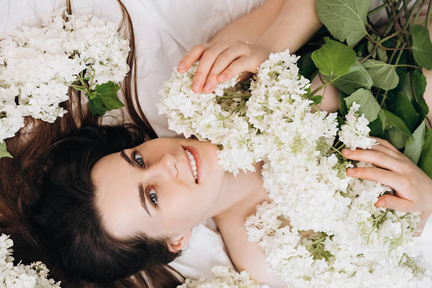 Top view portrait of beautiful smiling young woman with perfect skin, attractive dreamful caucasian female lying in cozy white bed near fresh flowers, happy looking at camera, natural beauty concept - Foto, Imagen
