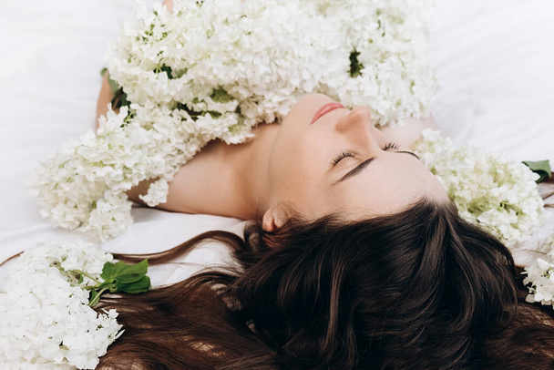 Romantic close up portrait of charming millennial brunette woman lying on comfortable white bed near fresh white flowers. Top view of cute calm attractive young girl smelling fresh seasonal flowers - Fotó, kép
