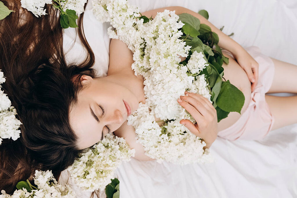 Sideview of cheerful beautiful pregnant young woman gently strokes tummy hand lying on cozy white bed at home with fresh bouquet flowers. Concept motherhood and maternity Future mother's expectation - Foto, imagen
