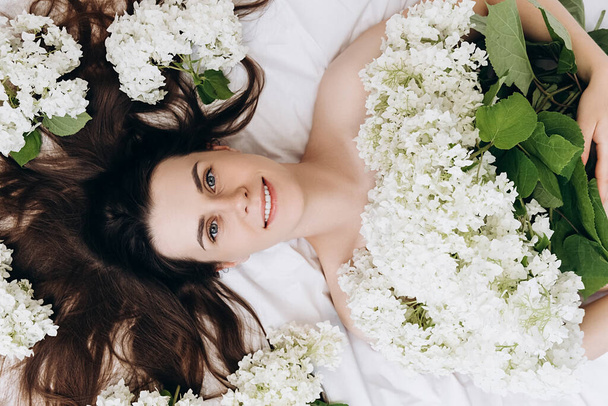 Top view portrait of smiling millennial young woman sincere happy looking at camera laying in cozy white bed, beautiful flowers around head. Cheerful brunette girl with clear skin enjoys, feels joy - Fotoğraf, Görsel