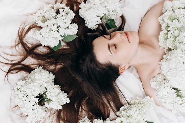 Top view beauty portrait of sexy brunette girl lying on comfy white bed near small beautiful flowers. Slender young woman, perfect clean skin and long hair. Natural cosmetics for skin care. - Foto, Bild