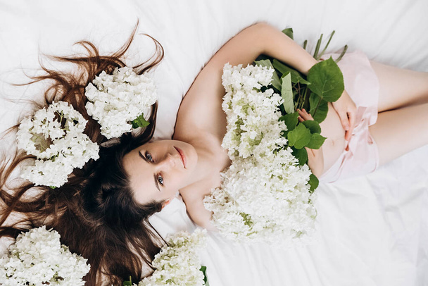 Side view close up portrait of cute millennial lady with fresh spring flowers, lying on comfy bed. Face of young cute caucasian woman with healthy clean skin. Natural face cosmetics, flower petals - 写真・画像