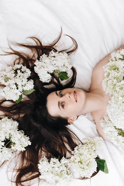 Close up portrait of happy millennial lady lying on cozy white bed near beautiful fresh spring flowers. Smiling girl enjoy glowing face skin after body care or treatment. Beauty treatment concept - Foto, Bild