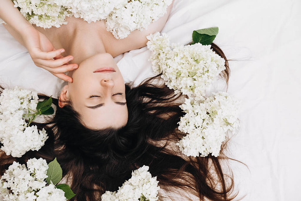 Top view of romantic smiling beautiful millennial lady with clean pure skin after beauty procedure, lying in bed with spring fresh white flowers. Skin beauty health care concept. Body part and nature - Foto, imagen