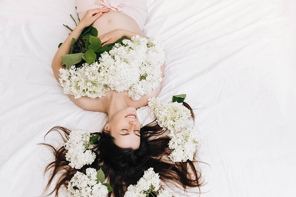 Piękna kobieta w ciąży z wiosennych lub letnich kwiatów trzyma ręce na brzuchu leżące na przytulnym białym łóżku w domu. Młoda kobieta czeka na narodziny dziecka. Ciąża, macierzyństwo, Dzień Matki Koncepcja wakacji - Zdjęcie, obraz