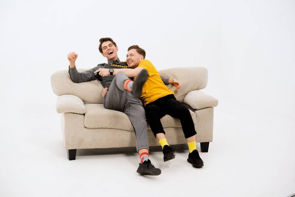 Funny friends on the white couch. Two guys on the couch laughing. Gay couple in love. Two young handsome boys sitting on a beige couch isolated on white background - Photo, Image