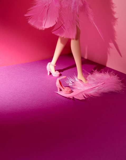 Kobieca lalka ubrana w różowe pióra i różowe buty na obcasie. Jasnoróżowe i fioletowe tło. Miejsce na tekst. Styl życia, moda, urok, koncepcja. Minimalny styl. - Zdjęcie, obraz