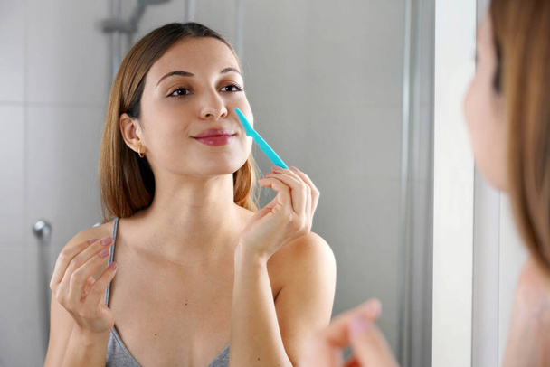 Hermosa mujer caucásica joven afeitándose la cara con navaja de afeitar en casa. Mujer bonita usando maquinilla de afeitar en baño. - Foto, Imagen