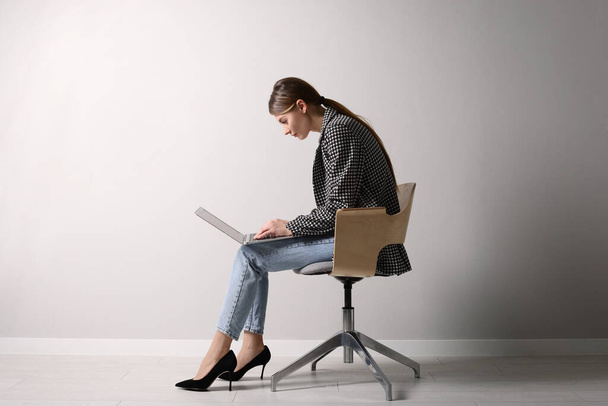 Woman with bad posture using laptop while sitting on chair near light grey wall indoors - Foto, imagen