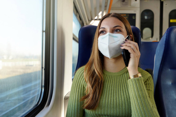 Jeune femme d'affaires portant un masque de protection requis pour les transports publics parlant avec un téléphone assis dans le train - Photo, image