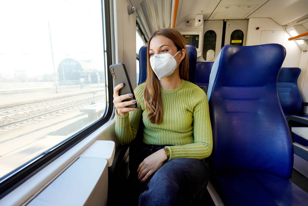 Νεαρή γυναίκα που φοράει ιατρική μάσκα χαλαρώνοντας στο κάθισμα του τρένου ενώ χρησιμοποιεί την εφαρμογή smartphone. Επιχειρηματίας απολαμβάνει προβολή γραπτών μηνυμάτων στο κινητό τηλέφωνο. Ασφάλεια ταξιδιού. - Φωτογραφία, εικόνα