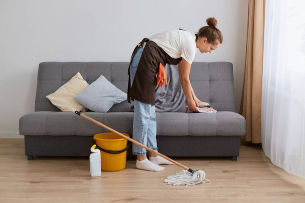 Side view image of young woman cleaning her home, wearing jeans and brown apron, housewife doing her household chores at home in light living room near window. - Foto, afbeelding