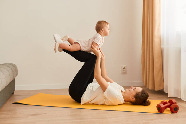 Side view of a woman in black leggings and a white T-shirt, a girl lying on the floor on a mat on her back with her legs raised up and holding a small child, home coach. - 写真・画像