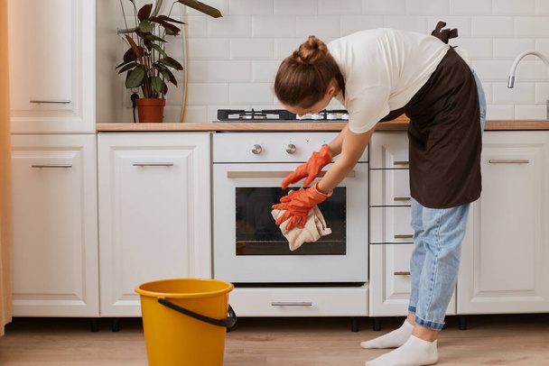 Indoor shot of woman washing steel gas stove and oven. Cleaning the apartment during her day-off, wearing jeans, white t shirt and brown apron, household chores. - Foto, Bild