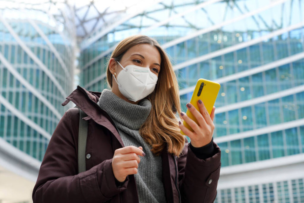 Επιχειρηματική γυναίκα που καλύπτει το πρόσωπο με προστατευτική μάσκα κρατώντας το κινητό τηλέφωνο με το σύγχρονο υπόβαθρο της πόλης. Τεχνολογία, ανάπτυξη, έννοια της υγειονομικής περίθαλψης. - Φωτογραφία, εικόνα