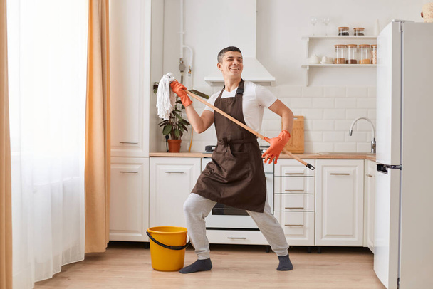 Full length portrait of positive optimistic dark haired Caucasian man wearing brown apron washing floor in kitchen having fun while cleaning house, dancing with mop. - Φωτογραφία, εικόνα