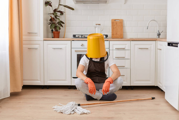 Portrait of unknown anonymous male housekeeper sitting on floor in kitchen with yellow bucket on his head, cleaning house, washing floor, being tired. - Foto, Imagen