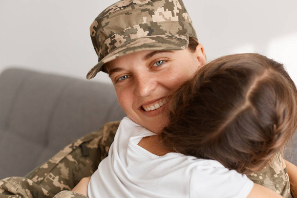 Portrait of soldier woman looking at camera with happy charming smile hugging her dark haired daughter, missing so much, returning home from army. - Foto, afbeelding