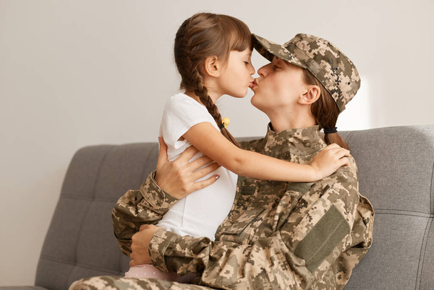 Indoor shot of happy military woman wearing camouflage uniform and cap, returning home from war or army, sitting on sofa with her daughter, kissing her child, missing, hugging each other. - Photo, image