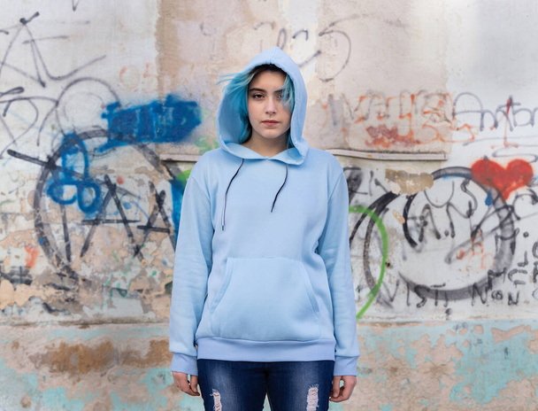 Adolescent portant un sweat à capuche oversize bleu clair et un jean regardant dans la caméra. Jeune fille aux cheveux bleus reste à l'extérieur contre le mur de graffiti. Modèle de vêtements  - Photo, image