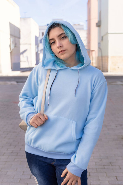 Teenager wearing light blue oversize hoodie and jeans looking into the camera. Blue haired teen girl stays outdoor on city street. Clothing mockup  - Φωτογραφία, εικόνα