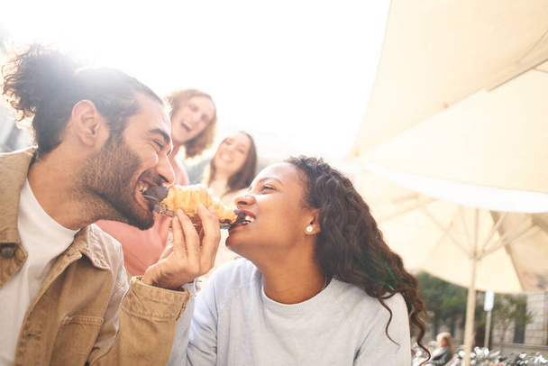 Foto divertente di una coppia che mangia un croissant come uno scherzo mentre gli amici guardano sullo sfondo. - Foto, immagini
