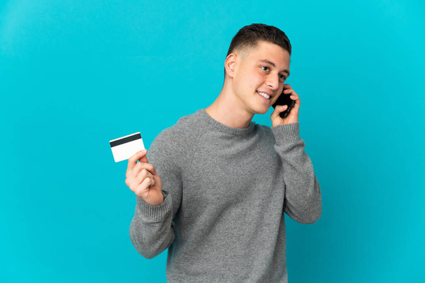 Joven hombre caucásico aislado sobre fondo azul manteniendo una conversación con el teléfono móvil y sosteniendo una tarjeta de crédito - Foto, imagen