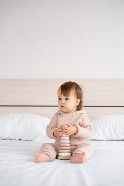 Екологічно чисті іграшки без пластику для немовлят. маленька дівчинка сидить вдома на ліжку і тримає сучасну дерев'яну барвисту піраміду з кільцями в руках. Місце для тексту
 - Фото, зображення