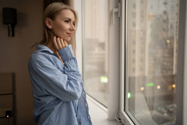 Portrait of a young pensive woman standing at the window in casual clothes getting ready for a special evening, copy space. - Photo, Image