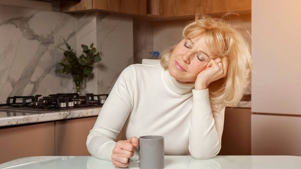Mulher de meia-idade sonolenta bebe café sentado na cozinha - Foto, Imagem