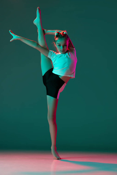 Little flexible girl, rhythmic gymnastics artist training isolated on green studio background in neon pink light. Grace in motion, action. Doing exercises in flexibility. - Photo, image