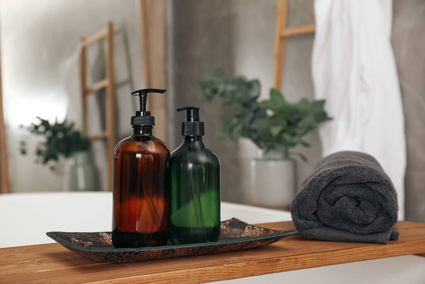 Wooden bath tray with bottles of shower gels and towel on tub indoors, space for text - Fotó, kép