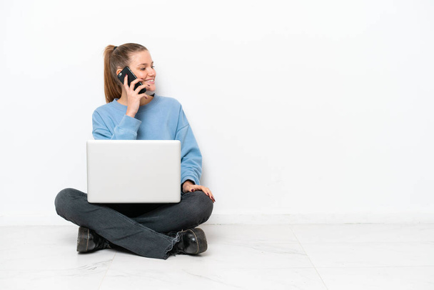 Dizüstü bilgisayarı olan genç bir kadın yerde oturuyor ve cep telefonuyla sohbet ediyor. - Fotoğraf, Görsel