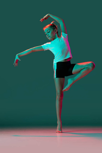 Dancing. Little flexible girl, rhythmic gymnastics artist training isolated on green studio background in neon pink light. Grace in motion, action. Doing exercises in flexibility. - Photo, image